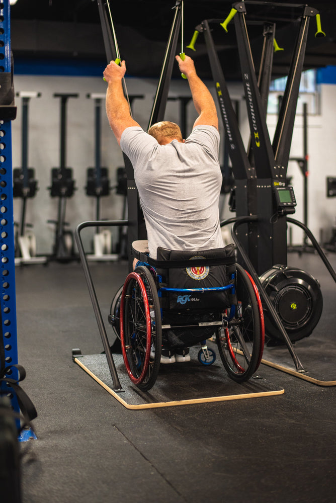 Pictured Kevin Ogar from the back using a Concept2 Ski Erg with the Equip Products Wider Base for Wheelchair users. In a gym setting on a black floor.