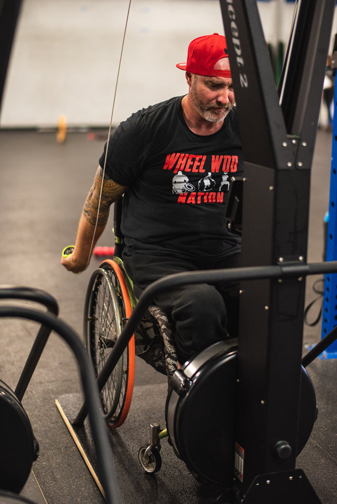 Chris "Stouty" Stoutenberg in his wheelchair and pulling the handles downward on the Equip Products wider base for the Concept2 Ski Erg, in a gym setting on a black floor.