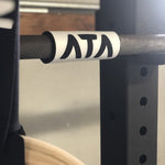 A white barbell marker on a barbell sitting in a rig. The barbell marker has the letters in the ATA logo printed in black.